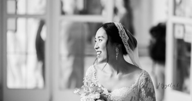 Chân dung cô dâu Châu Á mỉm cười trong trang phục cưới với voan viền ren và bông tai lấp lánh tại một địa điểm lịch sử ở Salzburg.